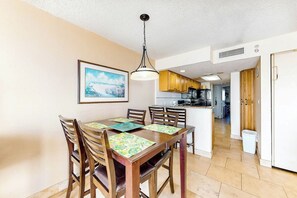 A dining area with a wooden table and chairs, a hanging light fixture, and a kitchen in the background. - Bay Watch Resort 0819 (North Myrtle Beach)