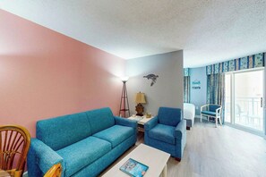 Ein Wohnzimmer mit einem blauen Sofa, einem Holzstuhl, einem weißen Couchtisch und einer Lampe. Es gibt ein Fenster mit Vorhängen und einen Blick nach draußen.