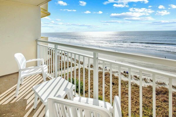 A balcony with white plastic chairs and a table, overlooking a beach with a fence and sparse vegetation. - Bay Watch Resort 0906 (North Myrtle Beach)
