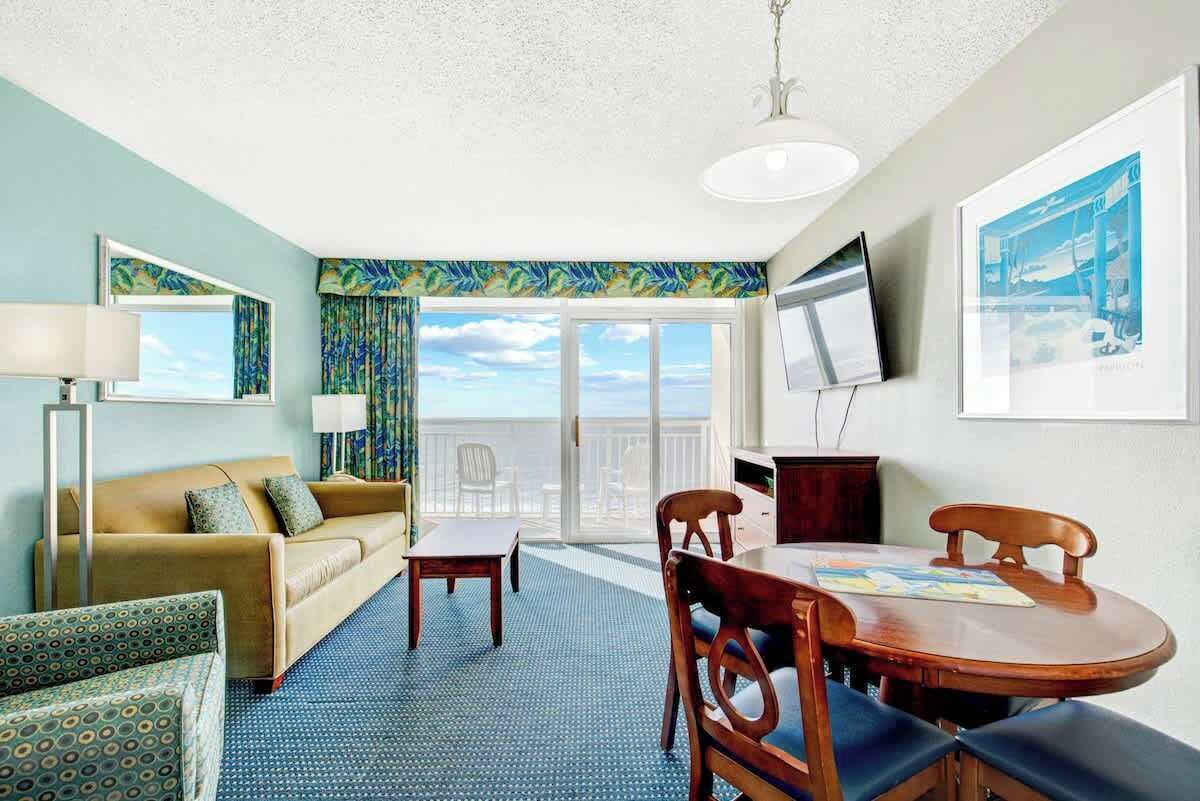 Habitación de hotel con sofá, una mesita, mesa de comedor, sillas, televisor y balcón con vistas.