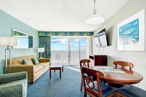 A hotel room with a sofa, a small table, a dining table, chairs, a TV, and a balcony with a view. - Bay Watch Resort 0906 (North Myrtle Beach)