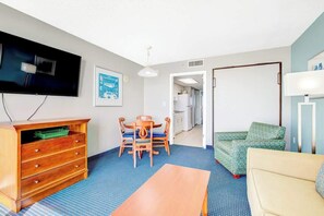 A compact living space with a television, a wooden dresser, a dining table with chairs, a sofa, and a patterned floor. - Bay Watch Resort 0906 (North Myrtle Beach)