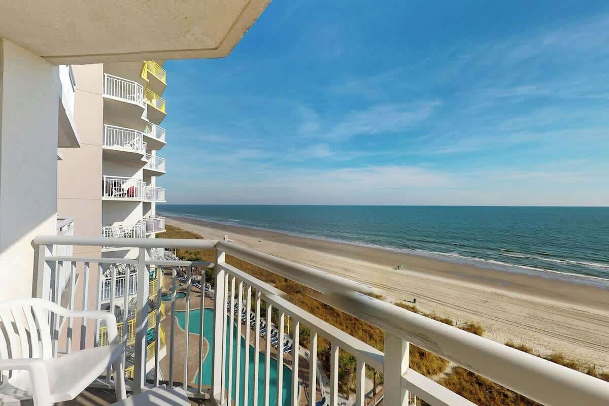 A balcony with a view of the beach and ocean, a pool, and white outdoor furniture.