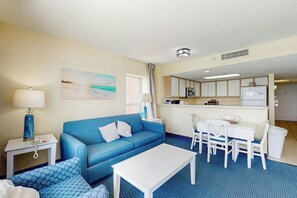 Ein Hotelzimmer mit einem blauen Sofa, einem weißen Couchtisch, einem Esstisch mit Stühlen, einer Küchenzeile und einem Bild an der Wand.