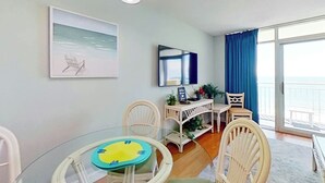 Ein Zimmer mit Glastisch, weißen Stühlen, einem Schreibtisch mit Computer, einem Sessel, einer Pflanze und Meerblick durch ein großes Fenster.