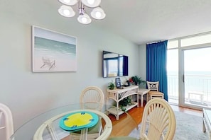 Ein Zimmer mit Glastisch, weißen Stühlen, einem Schreibtisch mit Computer, einem Sessel, einer Pflanze und Meerblick durch ein großes Fenster.
