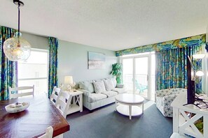 A living room with a dining table, sofa, and chairs. There is a window with curtains, a plant, and a painting on the wall. - Bay Watch Resort 1002 (North Myrtle Beach)