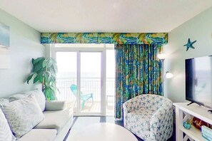 Ein Wohnzimmer mit einer Couch, einem Sessel, einem Fernseher und einem Balkon mit Vorhängen.
