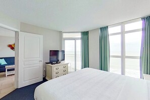 Habitación de hotel con cama, televisor en una cómoda, puerta y vista al océano a través de amplias ventanas.