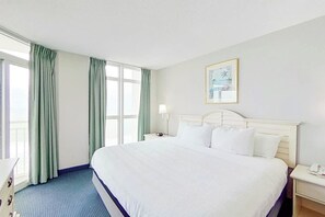 A hotel room with a large bed, a nightstand, a lamp, and a window with curtains. - Bay Watch Resort 1037 (North Myrtle Beach)