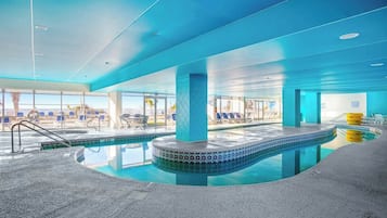 An indoor swimming pool with a blue-tiled edge, lounge chairs, and a view of the outdoors.