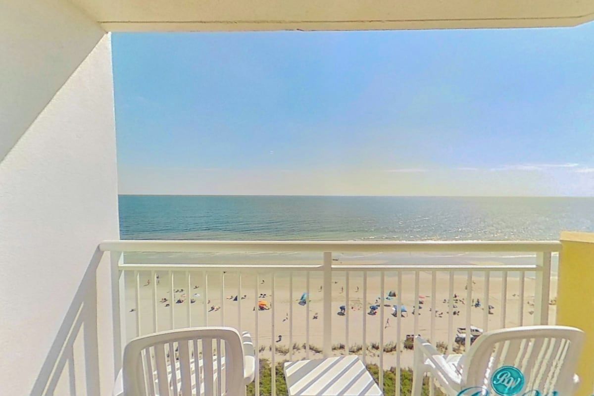 A balcony with white railings, two white chairs, and a table with striped tablecloths.