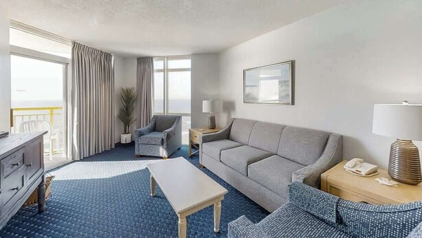 Quarto de hotel com uma janela grande, um sofá, uma cadeira, uma mesinha e uma mesa de cabeceira com abajur.