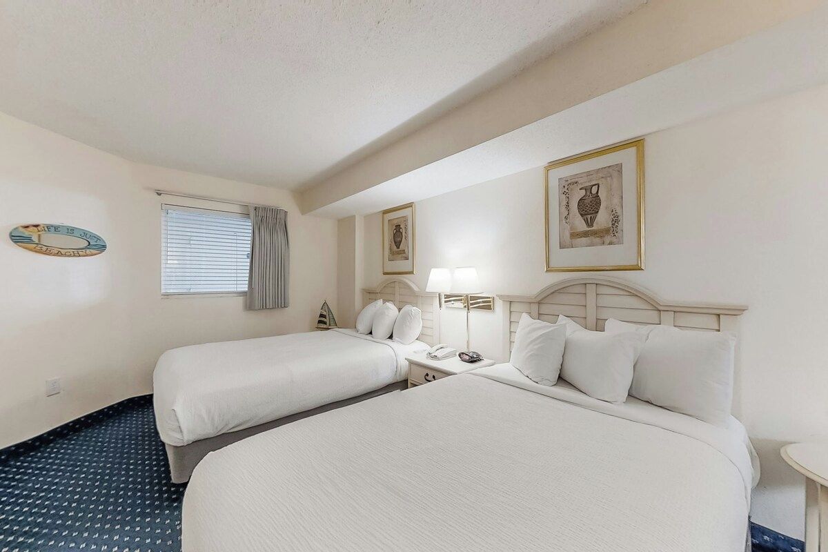 A hotel room with two beds, a bedside table with a lamp, a small table, and a window with blinds.