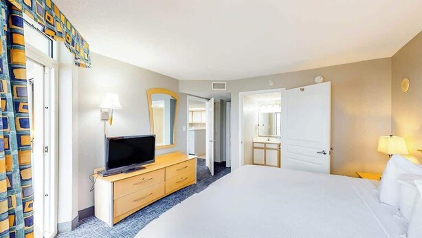 Ein Hotelzimmer mit einem Bett, einem Fernseher auf einer Holzkommode, einem Spiegel und einer Tür zu einem weiteren Raum.