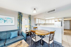 A modern living room with a sofa, a dining table with chairs, a kitchen area with cabinets, and a large window. - Bay Watch Resort 1209 (North Myrtle Beach)