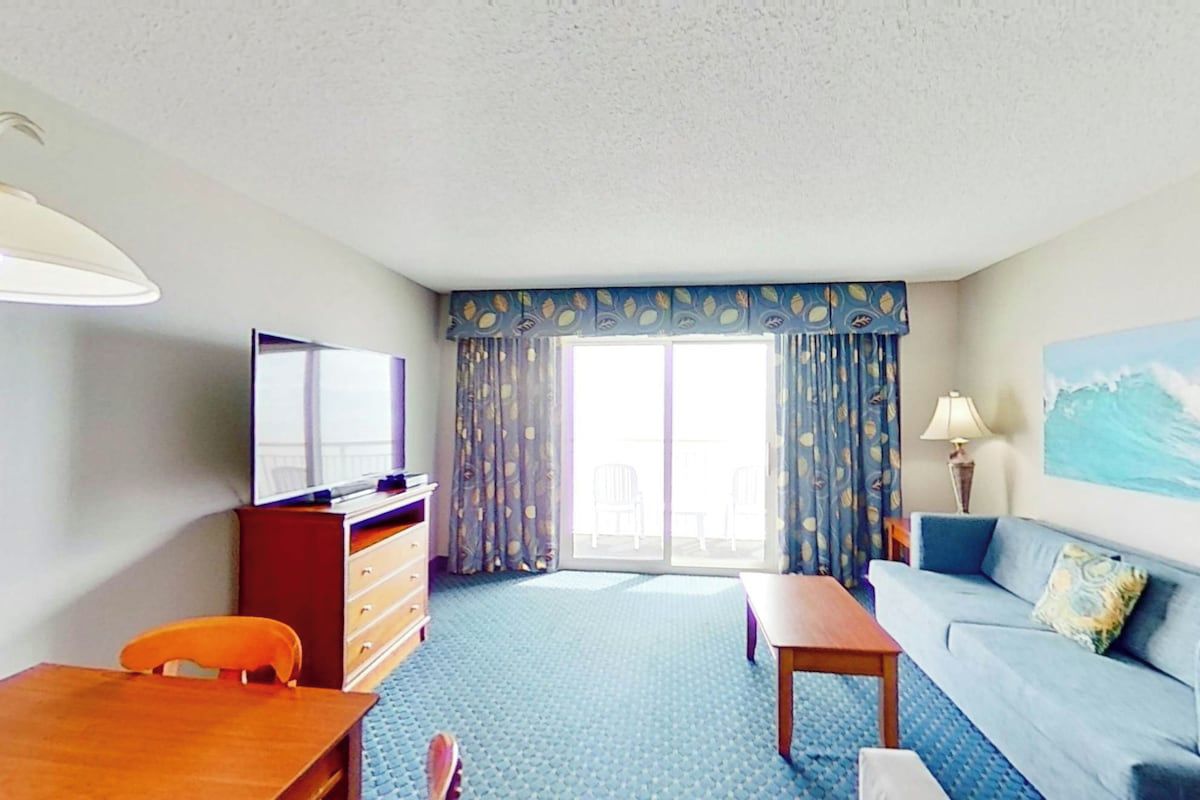 A hotel room with a television, a sofa, a table, a chair, a desk, and a large window with curtains.