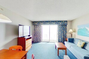 A hotel room with a television, a sofa, a table, a chair, a desk, and a large window with curtains. - Bay Watch Resort 1214 (North Myrtle Beach)