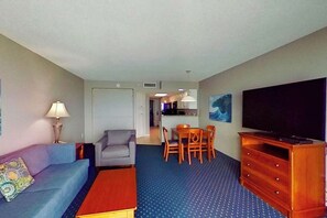 Ein Hotelzimmer mit einer Couch, einem Couchtisch, einer TV-Konsole, einem Essbereich mit Stühlen und einer Küche im Hintergrund.