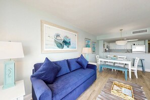 Sala de estar com sofá azul, mesa de jantar e cozinha com armários brancos e geladeira.