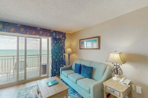 Ein Zimmer mit einer Couch, einem kleinen Tisch, einer Lampe, einem Telefon und Blick auf den Strand.