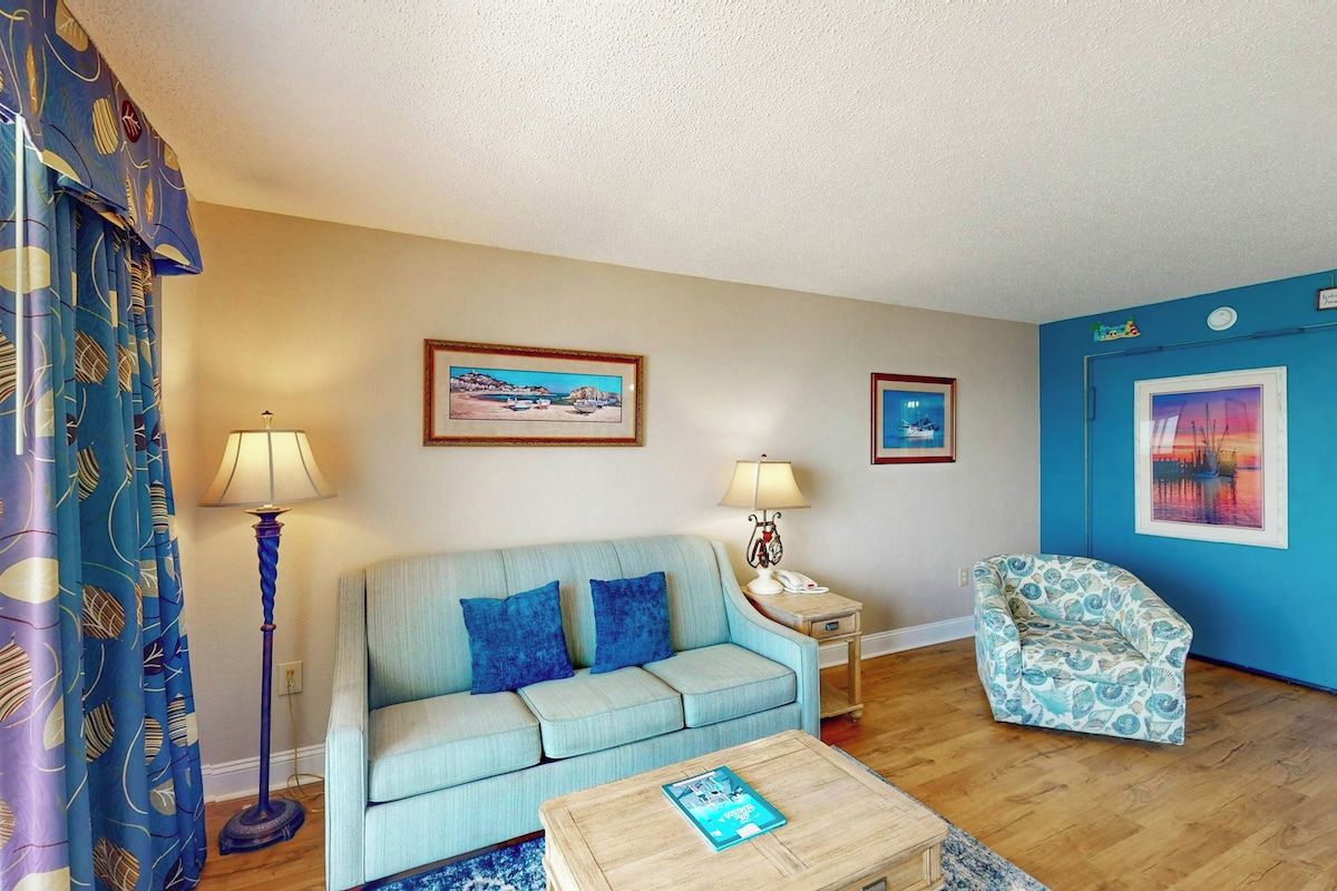 A living room with a sofa, armchair, coffee table, and two lamps. There are framed pictures on the walls.