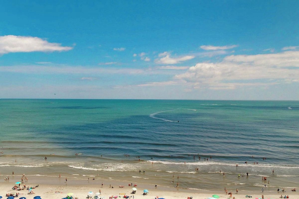 Uma praia com pessoas e guarda-sóis, céu claro e mar calmo.