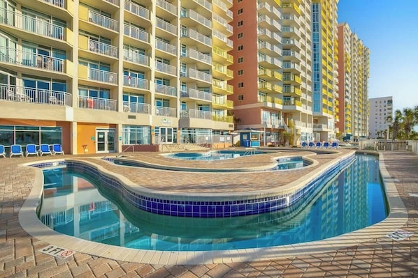Ein Hotel mit mehreren Etagen, einem Schwimmbecken mit Fliesenrand und blauem Himmel.