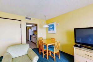 A compact room with a dining table, chairs, a sofa, a television, and a kitchen area in the background. - Bay Watch Resort 1104 (North Myrtle Beach)