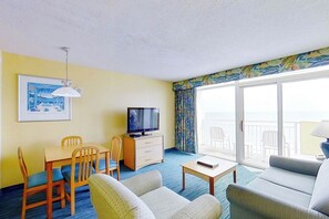A hotel room with a television, two sofas, a dining table with chairs, and a balcony with a view. - Bay Watch Resort 1104 (North Myrtle Beach)