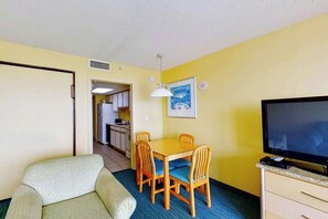 A compact room with a dining table, chairs, a sofa, a television, and a kitchen in the background. - Bay Watch Resort 1104 (North Myrtle Beach)