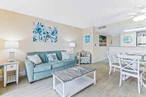 A living room with a sofa, coffee table, and a dining area with chairs. - Bay Watch Resort 1111 (North Myrtle Beach)