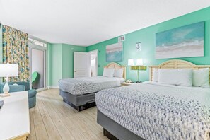 A hotel room with two beds, a desk, and a chair. There are two paintings on the wall, a door, and a window with curtains. - Bay Watch Resort 1111 (North Myrtle Beach)