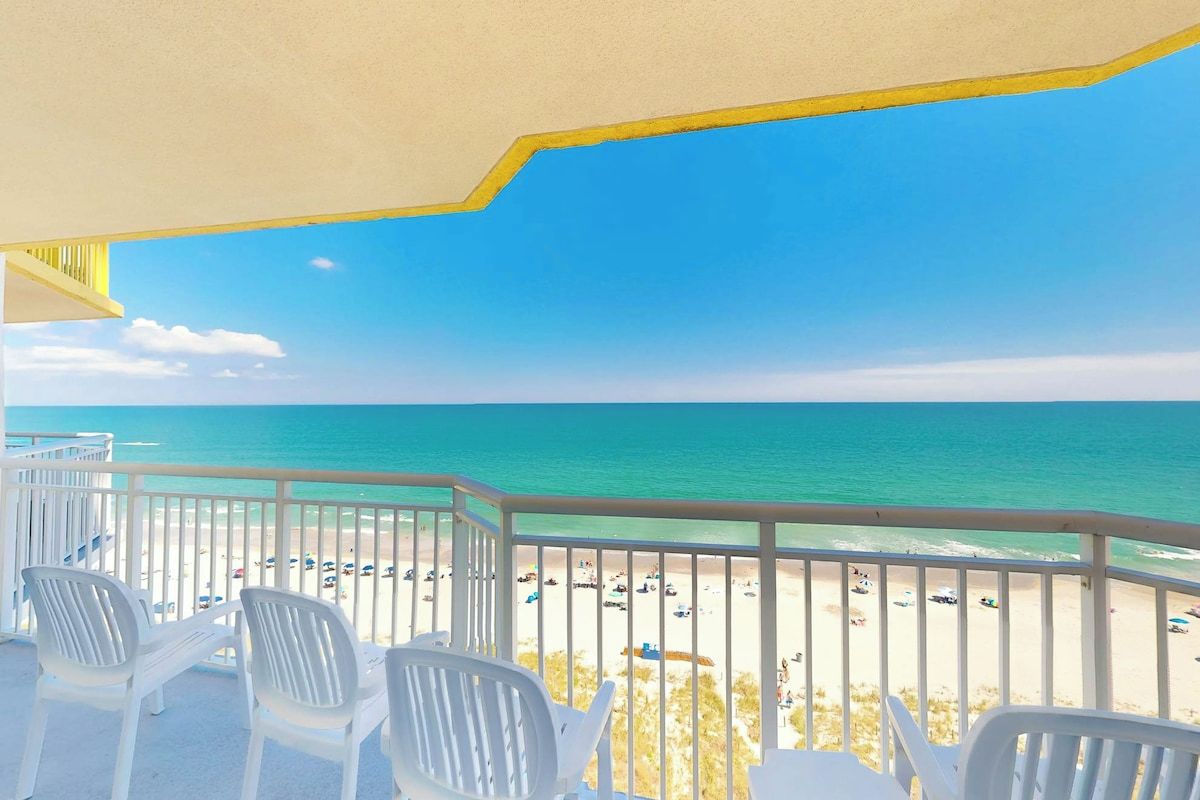 Ein Balkon mit weißen Plastikstühlen mit Blick auf einen Strand, an dem sich Menschen und Autos befinden.