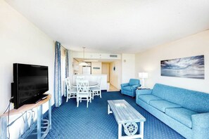 A hotel room with a TV, a dining table with chairs, a sofa, and a painting on the wall. - Bay Watch Resort 1022 (North Myrtle Beach)
