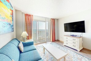 A room with a blue sofa, a white coffee table, a TV on the wall, and a sliding glass door leading to an outdoor area. - Bay Watch Resort 1238 (North Myrtle Beach)