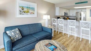 A living room with a sofa, a coffee table, a painting, and a kitchenette area. - Bay Watch Resort 1233 (North Myrtle Beach)