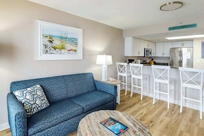 A living room with a sofa, a coffee table, a painting, and a kitchenette area. - Bay Watch Resort 1233 (North Myrtle Beach)