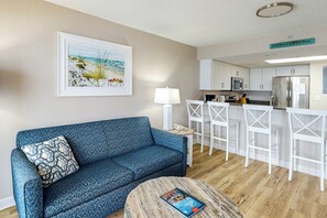 Ein Wohnzimmer mit einer Couch, einem Couchtisch mit einer Zeitschrift, einer Küchenzeile mit Kühlschrank, Mikrowelle und Barhocker.