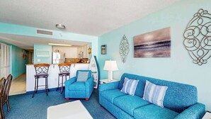 A living room with a blue sofa, a white coffee table, and a kitchen in the background. - Bay Watch Resort 1121 (North Myrtle Beach)