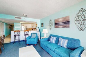 A living room with a blue sofa, a white coffee table, and a kitchen in the background. - Bay Watch Resort 1121 (North Myrtle Beach)