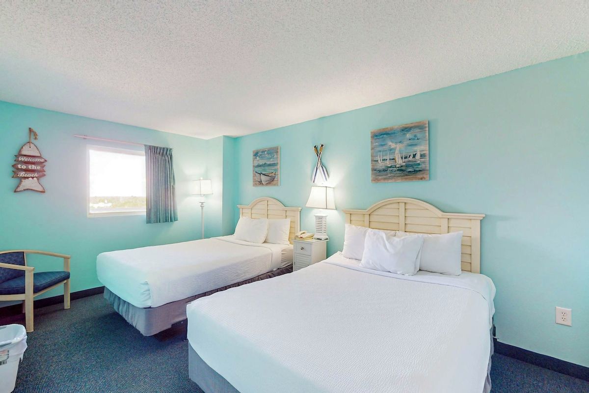 A hotel room with two beds, a chair, a nightstand, and a window with curtains.