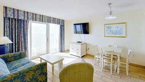 A living room with a sofa, a table, a TV, a dining set, and a window with curtains. - Bay Watch Resort 1135 (North Myrtle Beach)