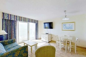 A living room with a sofa, a table, a TV, a dining set, and a window with curtains. - Bay Watch Resort 1135 (North Myrtle Beach)