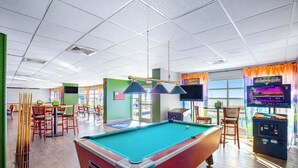 A pool table with a green felt surface, surrounded by wooden chairs and tables, with arcade machines and a television screen in the background. - Bay Watch Resort 1135 (North Myrtle Beach)