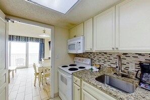A compact kitchen with a stove, microwave, and sink, leading to a dining area with a table and chairs. - Bay Watch Resort 1135 (North Myrtle Beach)