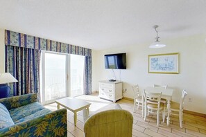 A living room with a sofa, a table, a TV, and a dining area with chairs. - Bay Watch Resort 1135 (North Myrtle Beach)