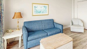 Ett rum med en mönstrad soffa, ett litet bord med en lampa, en inramad tavla på väggen och en stol.