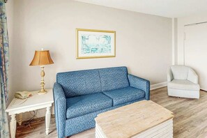 Ein Zimmer mit einem gemusterten Sofa, einem kleinen Tisch mit Lampe, einem gerahmten Bild an der Wand und einem Sessel.