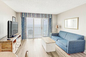 A living room with a television, a sofa, a coffee table, and a view through a glass door to the outside. - Bay Watch Resort 1439 Oceanfront (North Myrtle Beach)